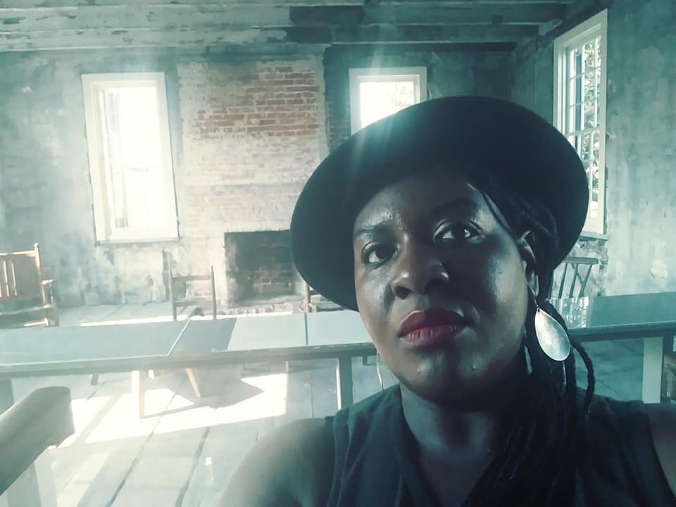 Photo of Dr. Moore in front of an old, empty room. She is glancing to the viewer's right, and wears a black hat and silver earrings.