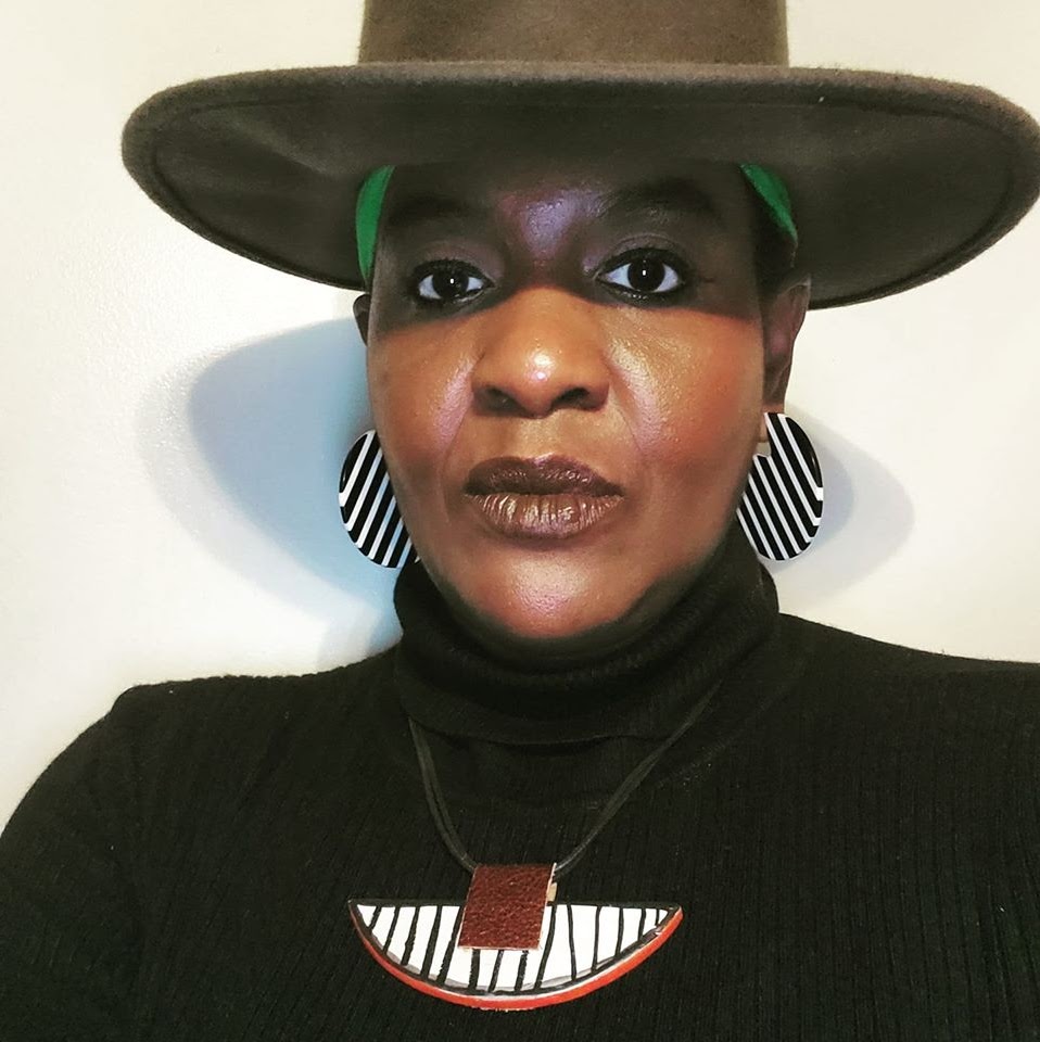 Portrait of Dr. Moore. She faces the camera against a white background, wearing a brown hat, black-and-white striped earrings and necklace, and a black turtleneck shirt.